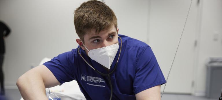 A man in a blue shirt assists another man lying in a hospital bed, providing care and support in a medical setting.