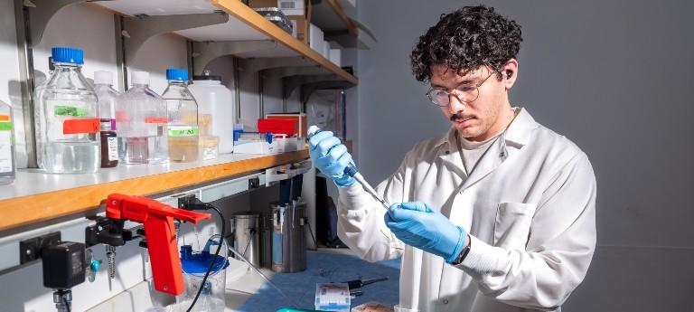 A student in a lab setting wearing protective equipment while working with material in a pipette