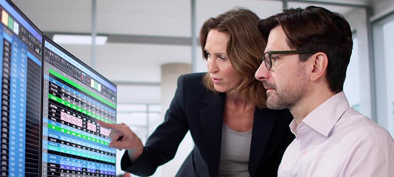 Two colleagues, a woman and a man, analyzing medical or scheduling data on a multi-monitor computer setup in an office.
