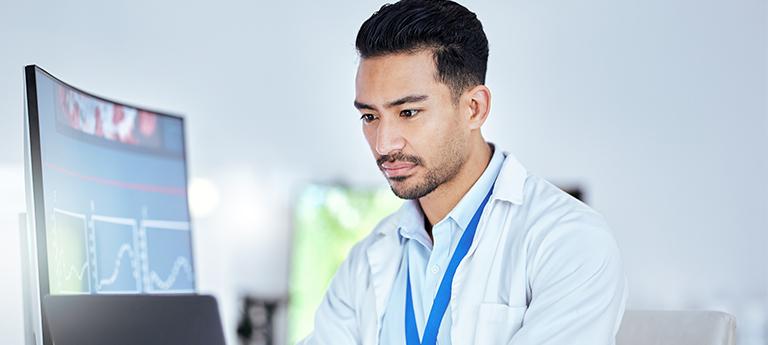  Male scientist or doctor in a lab coat concentrating on data visualizations and graphs on a computer monitor.