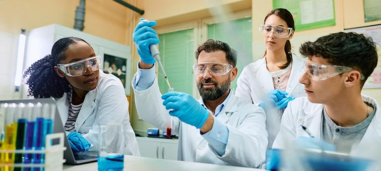Four researchers in lab coats and safety goggles collaborating on an experiment in a biochemistry laboratory.