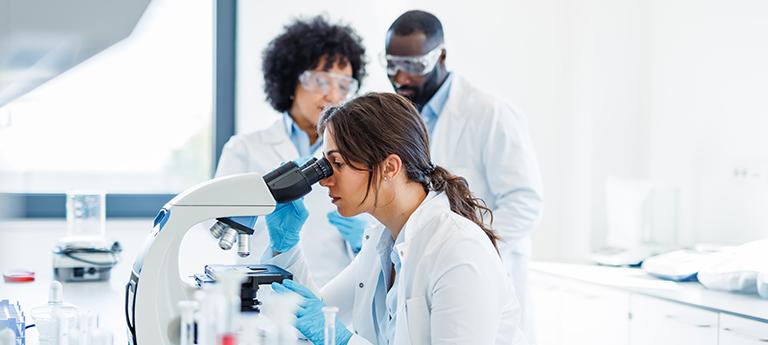 A female researcher looking through a microscope while two colleagues observe in a bright laboratory.