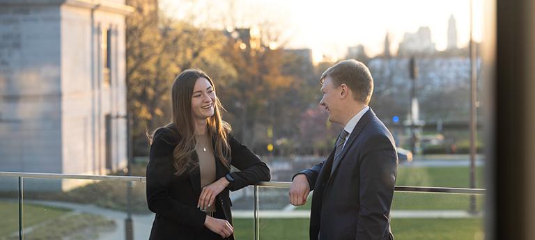 Two law students in professional attire laughing and talking on an outdoor balcony at sunset, with a view of the city skyline in the distance.