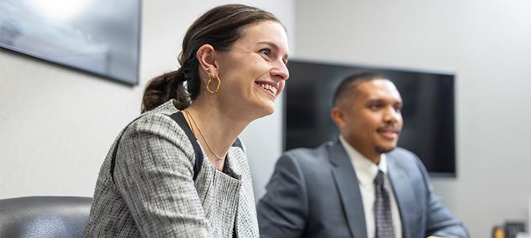 A close-up of a smiling woman in a grey patterned blazer participating in a discussion, with a colleague in a suit visible in the background of a modern office or classroom.