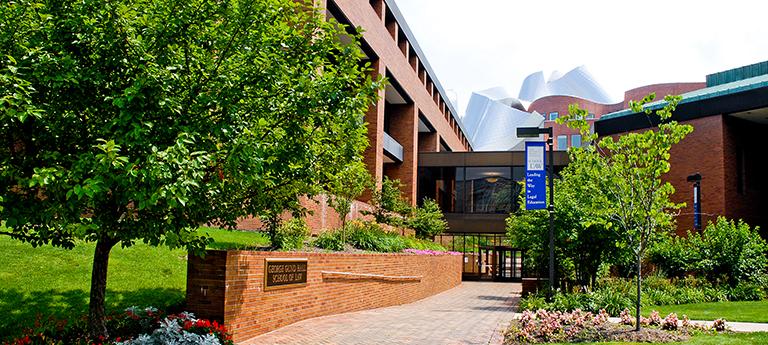 The George Gund Hall School of Law entrance, featuring a brick walkway, lush green trees, and the modern, metallic curves of the Peter B. Lewis Building in the background.