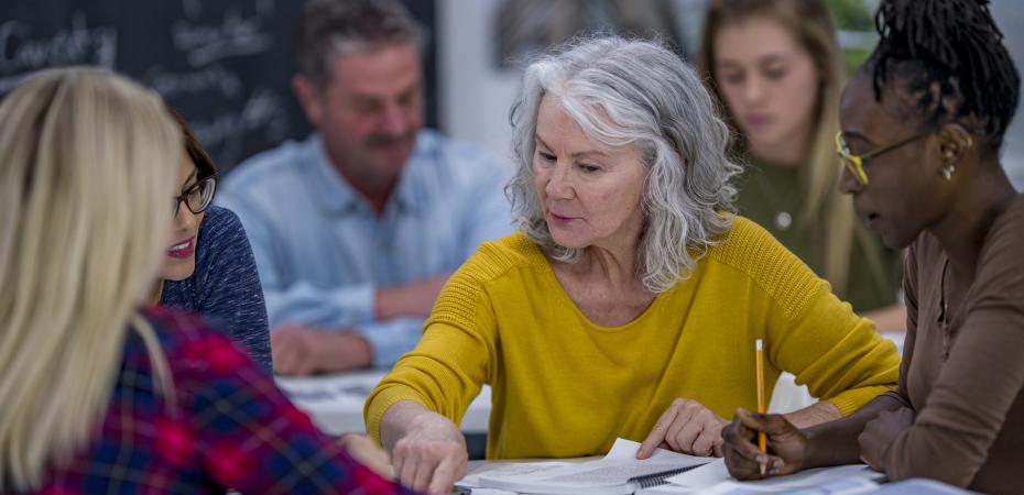 A group of university students are indoors in a study hall. They are having a group discussion. A Caucasian senior woman is reading out loud from a textbook.