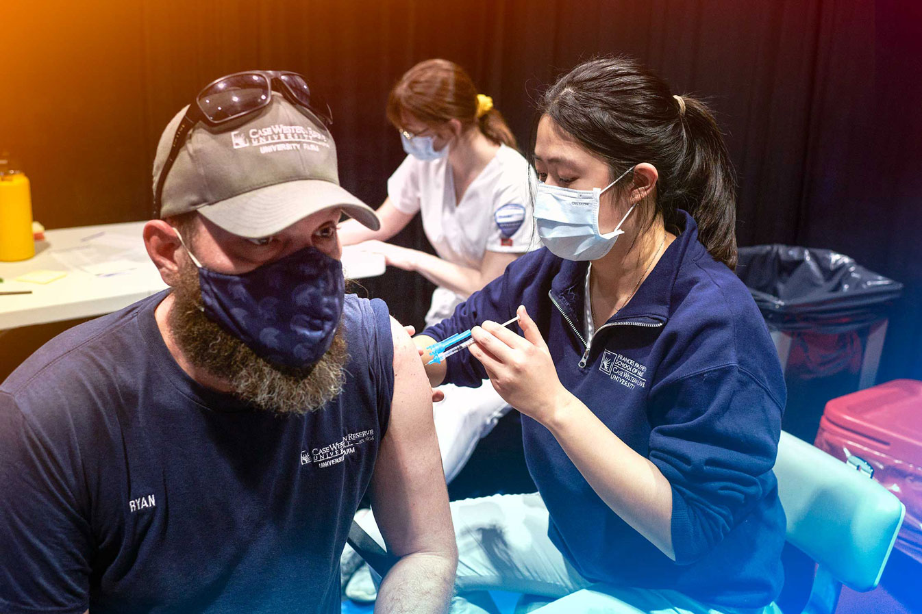 A woman wearing a mask administers a COVID-19 vaccine to a masked man while a nurse sits in the background at a CWRU vaccination clinic.