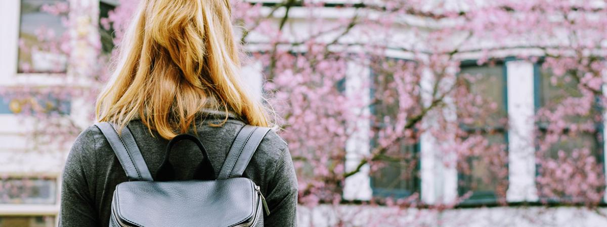A girl with a black backpack facing a blooming tree and building.