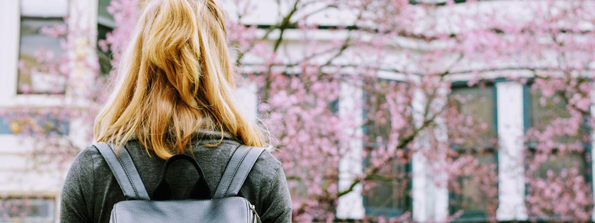 A girl with a black backpack facing a blooming tree and building.