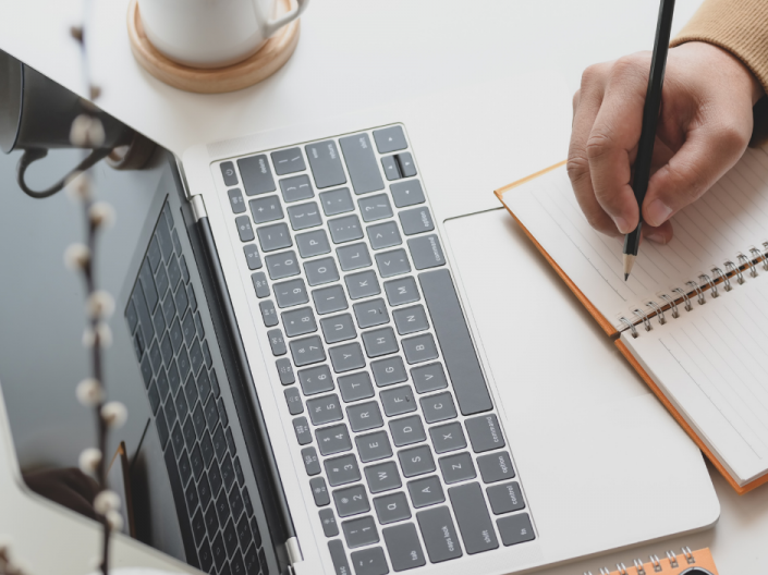 a person writes in a notebook while working on a laptop