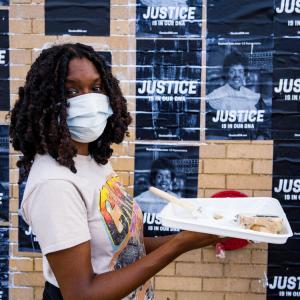 Photograph of a young Black woman gluing posters that read "Resistance is our DNA" onto the side of a building