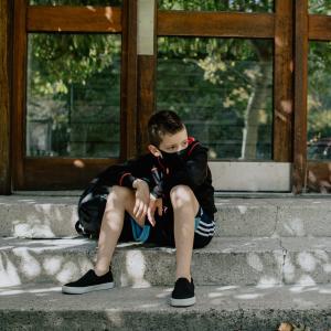 Photograph of a boy sitting on a curb looking pensive and wearing a mask to protect himself from COVID