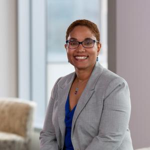 Photograph of Arielle Sheftall, a Black woman with closely cropped hair and glasses, wearing a gray suit and smiling at the camera