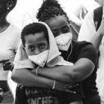 Photograph of young Black boy wearing a face mask being embraced from behind by his mother, also wearing a face mask, surrounded by protestors