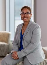 Photograph of Arielle Sheftall, a Black woman with closely cropped hair and glasses, wearing a gray suit and smiling at the camera