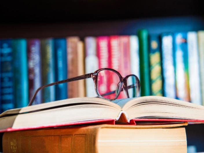 A pair of glasses sit on top of an opened book with a full bookshelf behind it