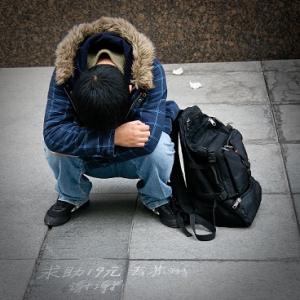 Man bent down and tucked into his knees wearing winter clothing and a book bag