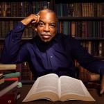 Photograph of actor and author LeVar Burton, seated at a table with a pile of books in front of him and filled bookcases behind him