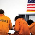 Young men in orange prison jumpsuits sitting with in a detention facility with their backs to the camera - an American flag hangs above them.