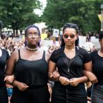 Photograph of four Black teenage girls wearing black clothes, with arms linked and fists raised at a Black Lives Matter protest