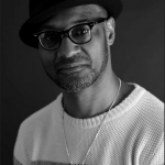 Photograph of Daniel Gray Kontar, a Black man wearing glasses and a hat, looking into the camera