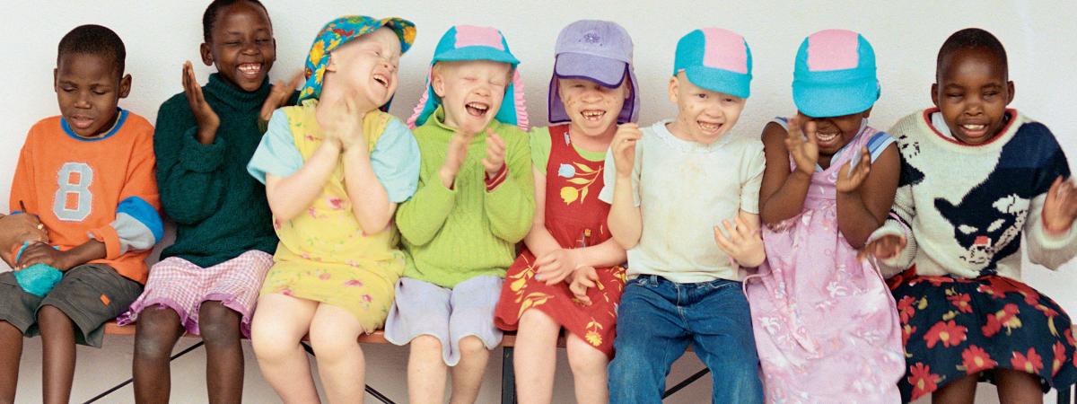 Photograph of laughing children sitting on bench in colorful clothes - some children are Black and some have light skin due to albinism. 