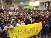 Photograph of a crowd of diverse school children in a library, holding a large banner that says "We need diverse books"