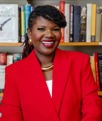 Kellie Carter Jackson standing in front of a bookshelf in a red blazer with white blouse underneath. Her braids swoop over the left side of her face, with the rest pulled back behind her
