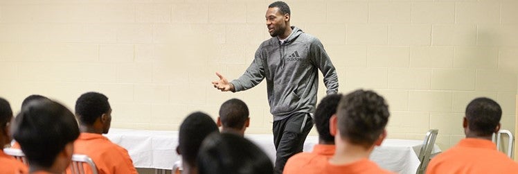 A man speaks to a group of youth wearing orange jumpsuits who appear to be in the juvenile justice system.