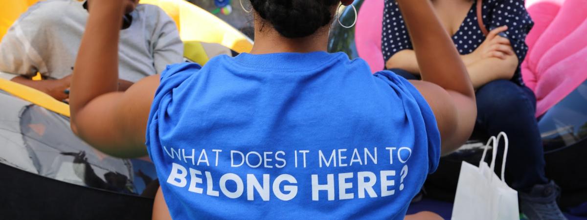Woman standing with her back to us wearing shirt with saying What does it mean to belong here
