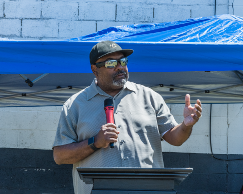 man wearing sunglasses talking into a microphone at an outdoor event