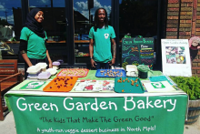 youth standing behind table of goods, tablecloth reads "green garden bakery, The kids that make the green good, a youth-run veggie dessert business in north mlps"