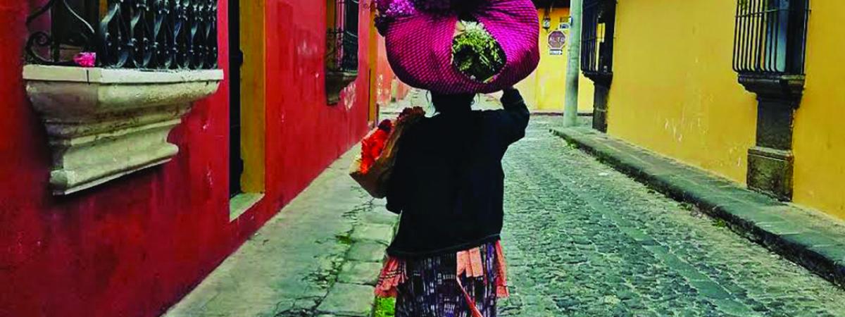 Image of woman in walking down an on old, but colorful, street in Guatemala, carrying bags in left arm and many bags on top of head