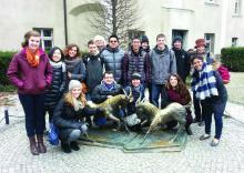 Image of sixteen students standing and crouching around a metal sculpture of two goats fighting, displayed on a city street paved with white-grey stones, with a white building and one bare tree in the background