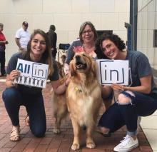 Social work students pose with a dog while holding All In signs on the steps of the Mandel School