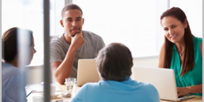 Image of four people sitting around a table, two with laptops, in a casual office setting