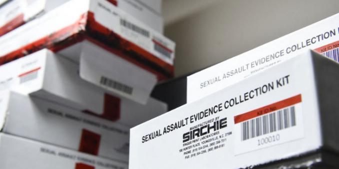Image of boxes of sexual assault evidence collection kits on a table, many stacked high on left, with two on right, the bottom one sticking out.  they are labeled sexual assault evidence collection kit manufactured by sirchie finger print laboratories 100 munter place, youngsville, n.c. 27596 phone 919-554-2244, 800-356-7311, fax 919-554-2266, 800-899-8181, with kit ik no 100010 on side of nearest box 