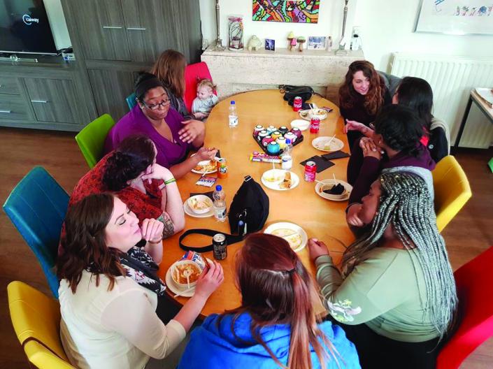 Image of nine students and baby sitting around large dinner table eating a meal in home with white and grey walls and creme colored fireplace with small items on mantel and abstract art in background