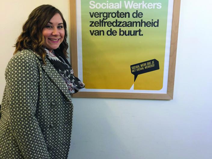 Student standing next to a poster that reads, Sociaal Werkers vergroten de zelfredzaamheid van de buurt, with a block caption that reads, sociaal werk doe je met sociaal werkers! below