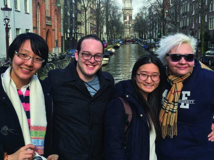 Image of four students on a bridge overlooking a canal with buildings and boats on each side, with a large cathedral spire in the background
