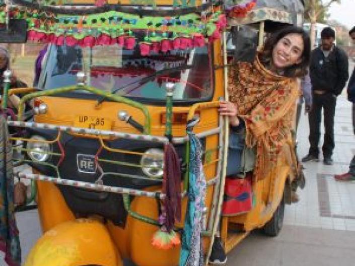 Image of woman in sari driving a yellow 3 wheel auto with very colorful scarves and decorations on it, down a street with a woman setting and two men standing with two trees in an open area