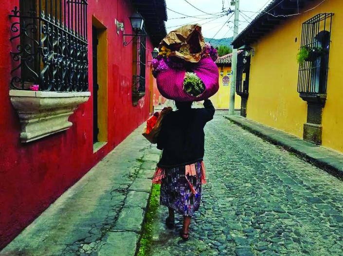 Image of woman in walking down an on old, but colorful, street in Guatemala, carrying bags in left arm and many bags on top of head