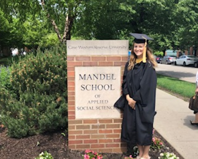 Rachel Truhan in graduation cap and gown standing in front of a sign for the Mandel School of Applied Social Sciences
