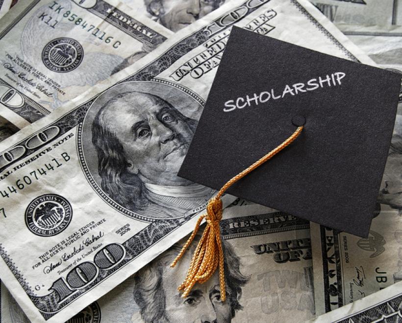 Mortar board cap with "Scholarship" written on it placed on top of $100 bills