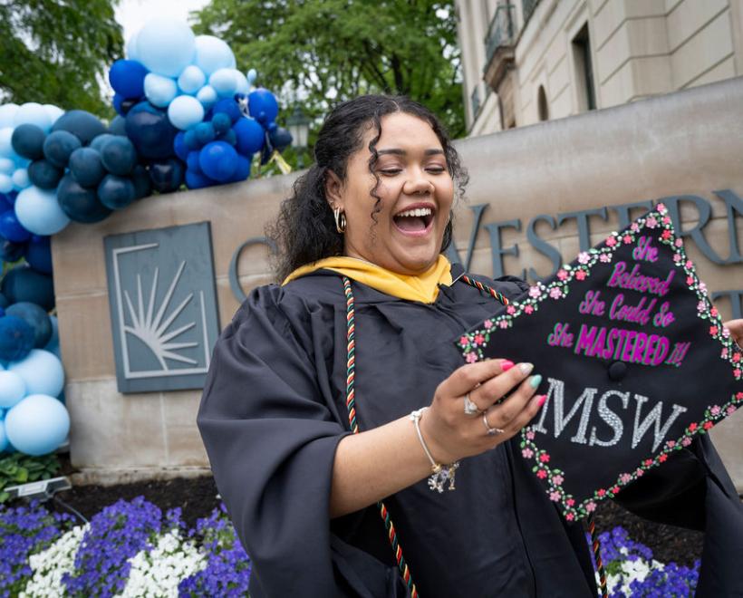 Woman holds her graduation cap in front of the CWRU sign on Euclid with balloons