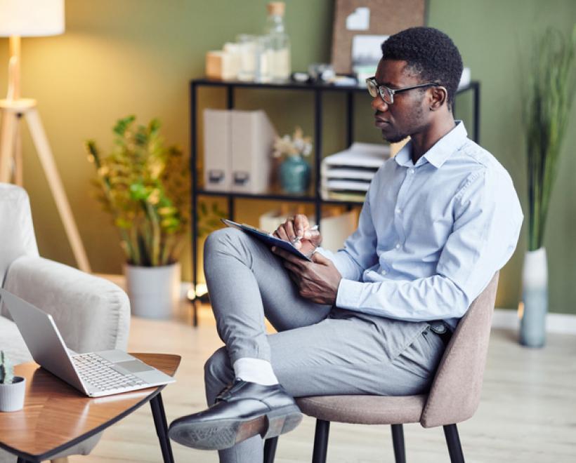 Person sits in a chair and takes notes while looking at a laptop on a table