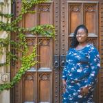 Tia Otoo standing in front of a big wooden door with vines crawling up it