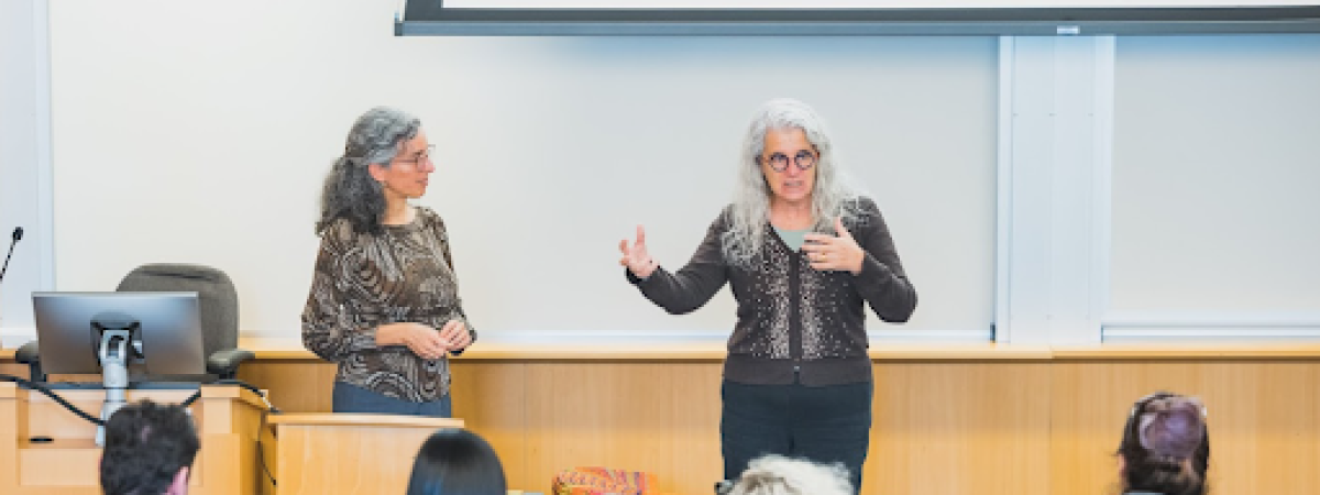 Two female presenting professors speaking to a class of students