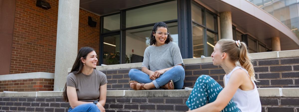 Three students sit on a set of wide stone stairs outdoors, talking to each other. 