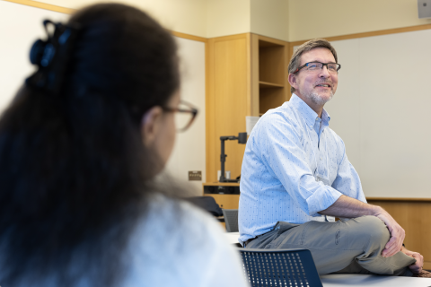 Smiling man sitting on a desk in a classroom appears to be talking to a student